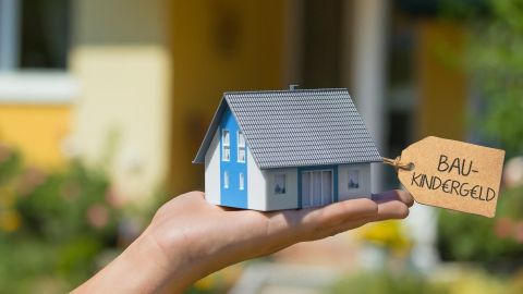 Eine Hand hält ein kleines Modellhaus mit einem Schild "Baukindergeld" vor einem unscharfen, hellen Hintergrund.