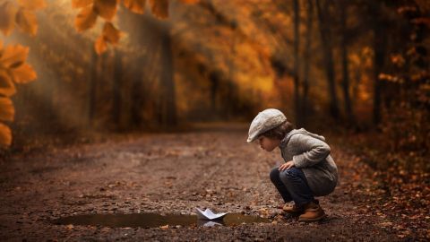 Ein Junge sitzt in einem herbstlichen Wald und beobachtet aufmerksam ein Papierboot im Pfützenwasser.