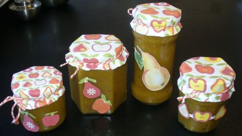 Vier dekorativ beschriftete Gläser mit fruchtigem Birnenchutney stehen auf einem dunklen Tisch in einer Küche.