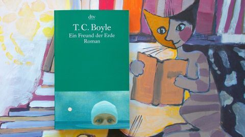 Ein grünes Buch von T.C. Boyle liegt auf einem Tisch, umgeben von einer bunten Malerei mit Katzen und einem Leser.
