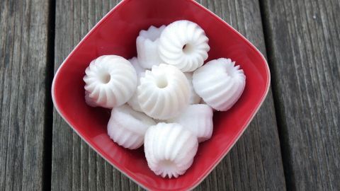 Ein rotes Schälchen enthält mehrere kleine, weiße Zucker-Gugelhupfformen, die auf einem dunklen Holzuntergrund arrangiert sind.