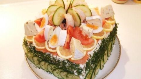 Eine üppige Smörgastarta steht auf einem weißen Teller, dekoriert mit Lachs, Gurken und Zitronen, in einer hellen Küche.