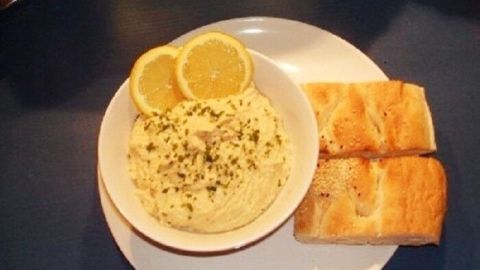 Eine Schüssel mit veganem Hummus, garniert mit Zitronenscheiben, liegt neben zwei Stückchen Brot auf einem Teller.