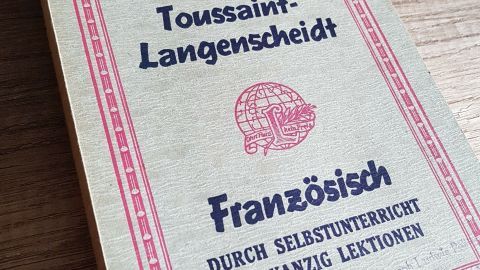 Eine alte Buchhülle liegt auf einem Holzuntergrund. Es handelt sich um ein Französisch-Lernbuch, das Gebrauchsspuren zeigt.