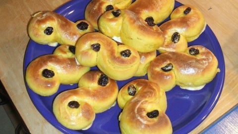 Auf einem blauen Teller liegen viele Lussekatter. Eine Figur im Hintergrund hat eine Krone und hält einen Tannenzweig.