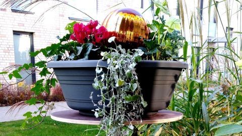Ein Sonnenschirmständer ist kreativ umfunktioniert und hält blühende Pflanzen in einem grünen Gartenbereich.