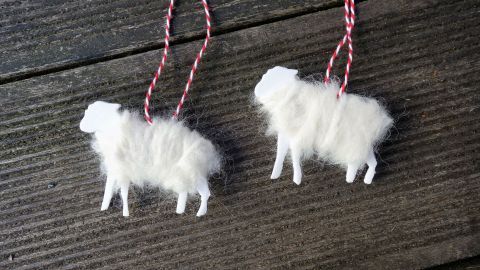 Zwei Weihnachts-Schafe aus weißen Materialien hängen an einer roten Schnur und sind auf einer dunklen Holzoberfläche platziert.