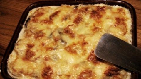 Ein goldbraunes Kartoffel-Pilz-Gratin wird aus einer Auflaufform mit einem Spatel serviert, umgeben von Holz.