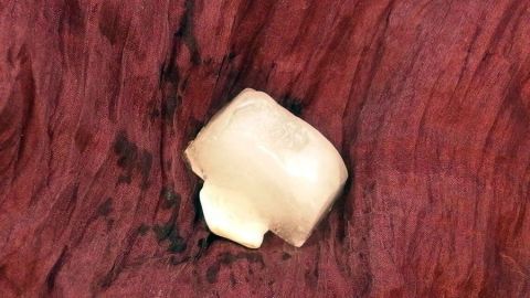 Ein Eiswürfel liegt auf einem roten Hintergrund, möglicherweise auf einem Tisch oder einer anderen Oberfläche, und reflektiert das Licht.