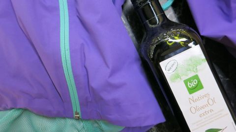 Mit etwas Spülmittel, Geduld und der richtigen Auswahl des Waschgangs entfernt man Ölflecken aus Goretex-Kleidung im Nu! Eine lila Goretex-Jacke neben einer Flasche Olivenöl, die einen Ölfleck auf dem Stoff hinterlässt.