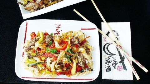 Ein asiatischer Rindfleischsalat liegt in einer weißen Schale neben einer Rindfleischpfanne auf einem dunklen Tisch.