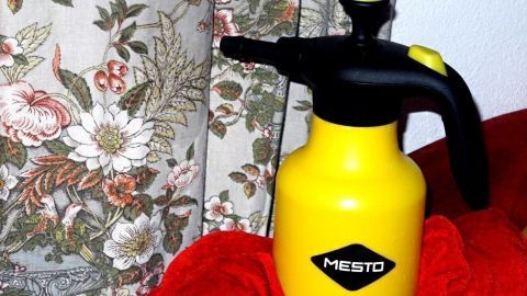 Ein gelbes Sprühgerät steht auf einem roten Tuch vor einem blumigen Vorhang, bereit, die Luftfeuchtigkeit in Wohnräumen zu erhöhen.
