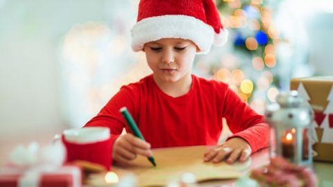 Ein Kind trägt einen Weihnachtsmann-Hut und schreibt konzentriert an einem Tisch mit Geschenken und Kerzen.