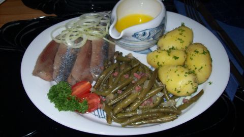 Ein Teller zeigt Matjesfilets neben Petersilienkartoffeln und grünen Speckbohnen, garniert mit Zwiebeln und Sauce.