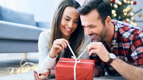Ein Paar öffnet ein rotes Geschenk unterm Weihnachtsbaum und freut sich über die Überraschung im festlichen Raum.