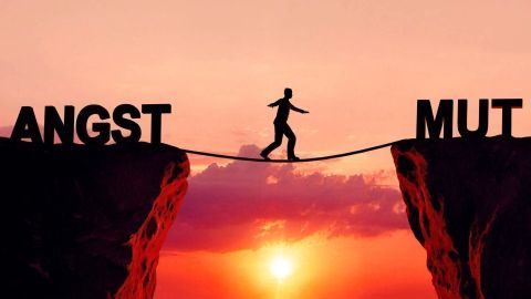 Eine Person balanciert auf einem Seil zwischen den Worten „Angst“ und „Mut“ vor einem Sonnenuntergang.