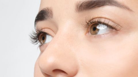 Lange, schön gebogene Wimpern umrahmen die Augen einer jungen Frau, die vor einem hellen, neutralen Hintergrund betrachtet werden.