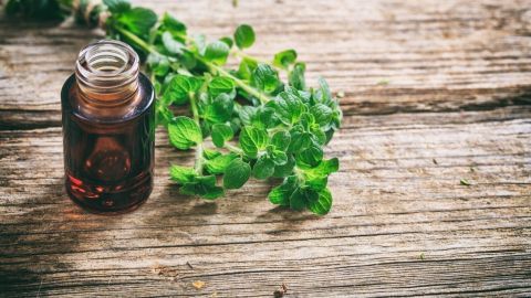 Ein braunes Fläschchen mit Oregano-Öl steht auf einer Holzoberfläche neben frischen Oregano-Blättern, als natürliche Lösung gegen Nagelpilz.