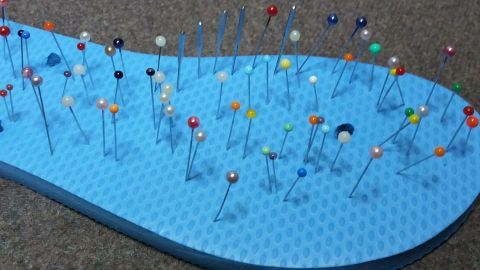 Eine blaue, strukturierte Unterlage hält viele bunte Nadeln aufrecht, übersichtlich angeordnet auf einem braunen Boden.