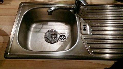 Eine Edelstahlspüle mit einem leicht sichtbaren braunen Belag steht zur Reinigung. Sie hat eine Abtropffläche und einen Wasserhahn.