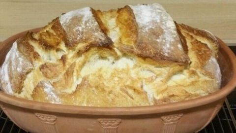 Ein frisch gebackenes Joghurtbrot ruht goldbraun und knusprig im Römertopf auf einem Ofengitter neben einer Holzfläche.