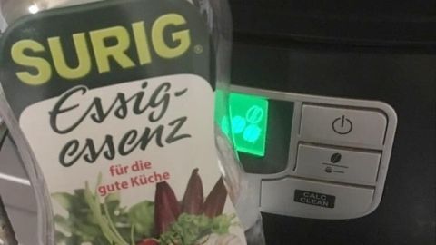 Eine Flasche Essig-Essenz steht neben einer Kaffeemaschine, die grün leuchtet, zum Entkalken und Reinigen.
