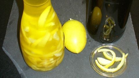 Ein Glas mit Zitronenschalenöl steht auf einem schwarzen Untergrund, neben einer Zitrone und einer dunklen Flasche.