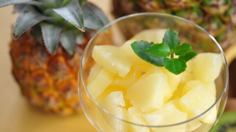 Ein Glas gefrorene Ananasstücke mit einem Minzblatt steht vor einer frischen Ananas auf einem hellen Tisch.