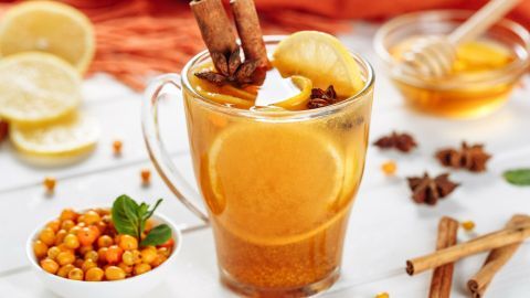Ein Glas Sanddorntee mit Ingwer und Zimt steht auf dem Tisch, umgeben von Zitrone, Honig und Beeren, in einem hellen Ambiente.