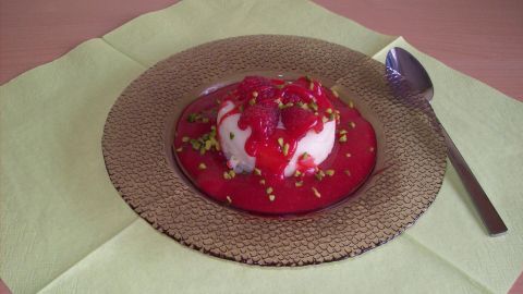 Pannacotta liegt auf einem goldenen Teller, überzogen mit Himbeermus und dekoriert mit Pistazien, auf einem grünen Tuch.
