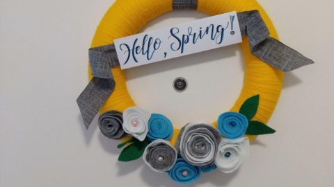 Ein gelber Türkranz mit bunten Papierblumen und einem Schild, das „Hello Spring!“ sagt, hängt vor einer weißen Wand.