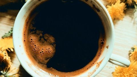 Eine Tasse Löwenzahnkaffee steht auf einem Holzuntergrund, umgeben von gelben Löwenzahnblüten. Der Kaffee zeigt eine dunkle, cremige Oberfläche.
