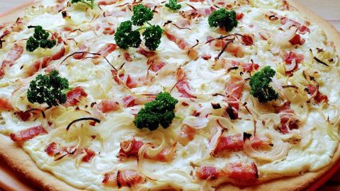 Ein frisch gebackener Flammkuchen liegt auf einem Holzbrett. Er ist belegt mit Speck, Zwiebeln und frischen Kräutern.