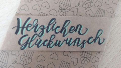 Eine Geschenkkarte ist auf einem beige-gestreiften Hintergrund platziert, darauf steht "Herzlichen Glückwunsch" in geschwungener Schrift.