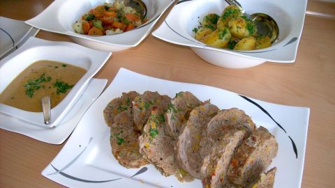 Saftiger Hackbraten aus dem Bratschlauch liegt auf einem Teller, umgeben von Beilagen in Schalen auf einem Tisch.
