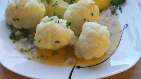 Blumenkohl in weißer Soße liegt auf einem Teller, umgeben von Salzkartoffeln, garniert mit frischer Petersilie.