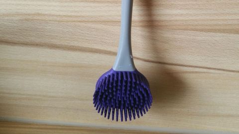 Eine Silikon-Spülbürste hängt an einer Wand, bereit, Geschirr zu reinigen, mit einem ergonomischen Griff in Grau und Lila.
