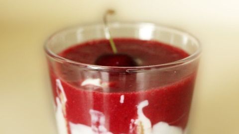 Eine gläserne Schüssel zeigt einen cremigen Nachtisch mit roten Fruchtsoße und einer Kirsche oben drauf, vor neutralem Hintergrund.