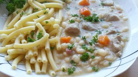 Eine Schale mit weißen Bohnen und zartem Schweinefleisch, umgeben von nudelartigen Beilagen und frischen Kräutern.