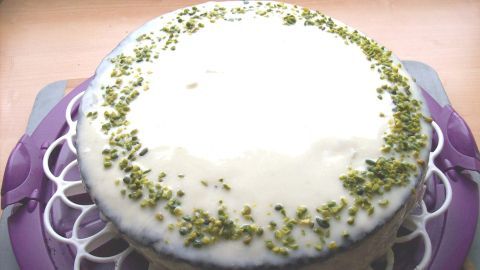 Ein Schoko-Kuchen mit Mirror Glaze liegt auf einem dekorativen Gitter auf einem Tisch, umrandet von grünen Pistazienstücken.