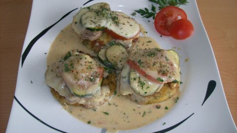 Medaillons vom Schwein liegen auf goldenen Kräuter-Kartoffel-Talern, serviert in einer cremigen Sauce mit Tomate.