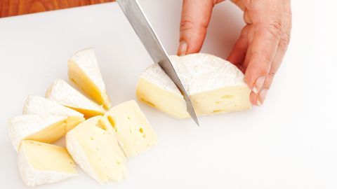Ein handgeschriebener Käse wird mit einem Messer auf einem Schneidebrett in gleichmäßige Stücke geschnitten.