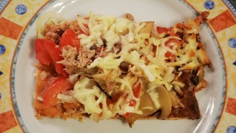 Ein Low Carb Pizzaboden liegt auf einem dekorativen Teller, belegt mit Käse, Gemüse und Fleisch, auf einer Arbeitsfläche.