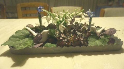 Eine Frühlingsschale mit bunten Blumen und Blättern schmückt einen Tisch in einem hellen Raum.