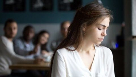 Eine nachdenkliche Frau sitzt in einem Café und wirkt emotional, während im Hintergrund andere Personen an einem Tisch plaudern.
