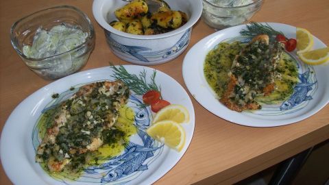 Zwei Teller mit Rotbarschfilet und Kräutertopping liegen auf einem Holztisch, umgeben von Beilagen und Dips.
