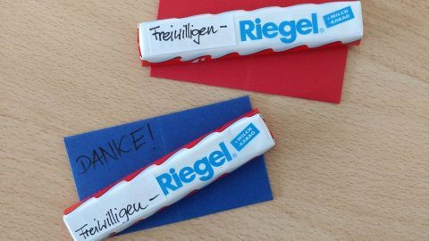 Ein roter und ein blauer kleiner Briefumschlag mit einer weißen Schokolade, darauf steht „Danke!“ und „Riegel!“, liegen auf einem Tisch.