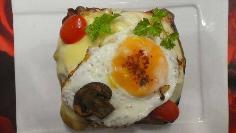 Ein überbackener Leberkäsetoast mit goldbraunem Spiegelei, Champignons und frischen Tomaten auf einem weißen Teller.