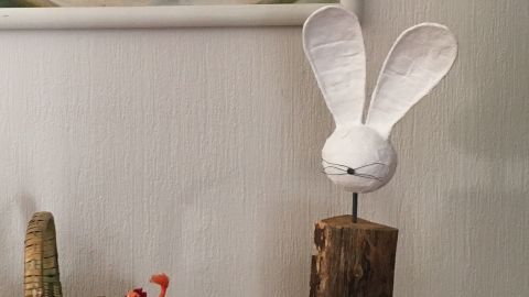 Ein weißer Osterhase aus Papier sitzt auf einem Holzstück in einem gemütlichen Wohnraum neben einem geflochtenen Korb.