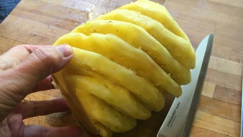 Eine Hand hält eine attraktiv geschnittene Ananas über einem Holzbrett, umgeben von einem scharfen Messer.